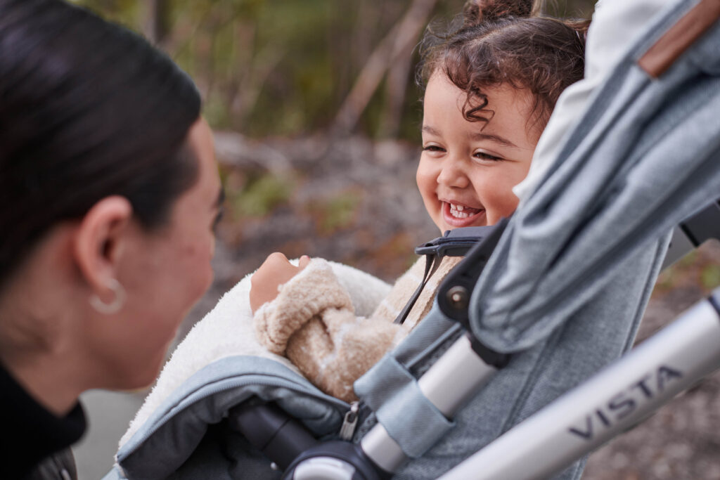 A child smiling at mom while snug in their UPPAbaby Vista V2 stroller.